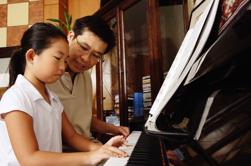 A young child seated at an upright piano with an encouraging parent nearby