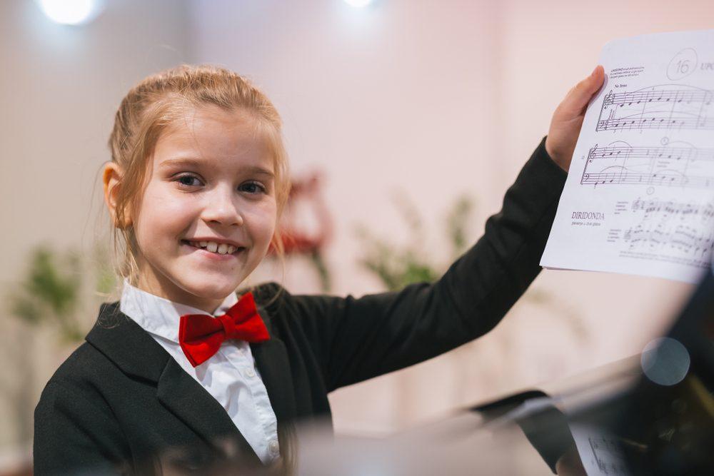A young piano student in recital attire smiling and holding her sheet music